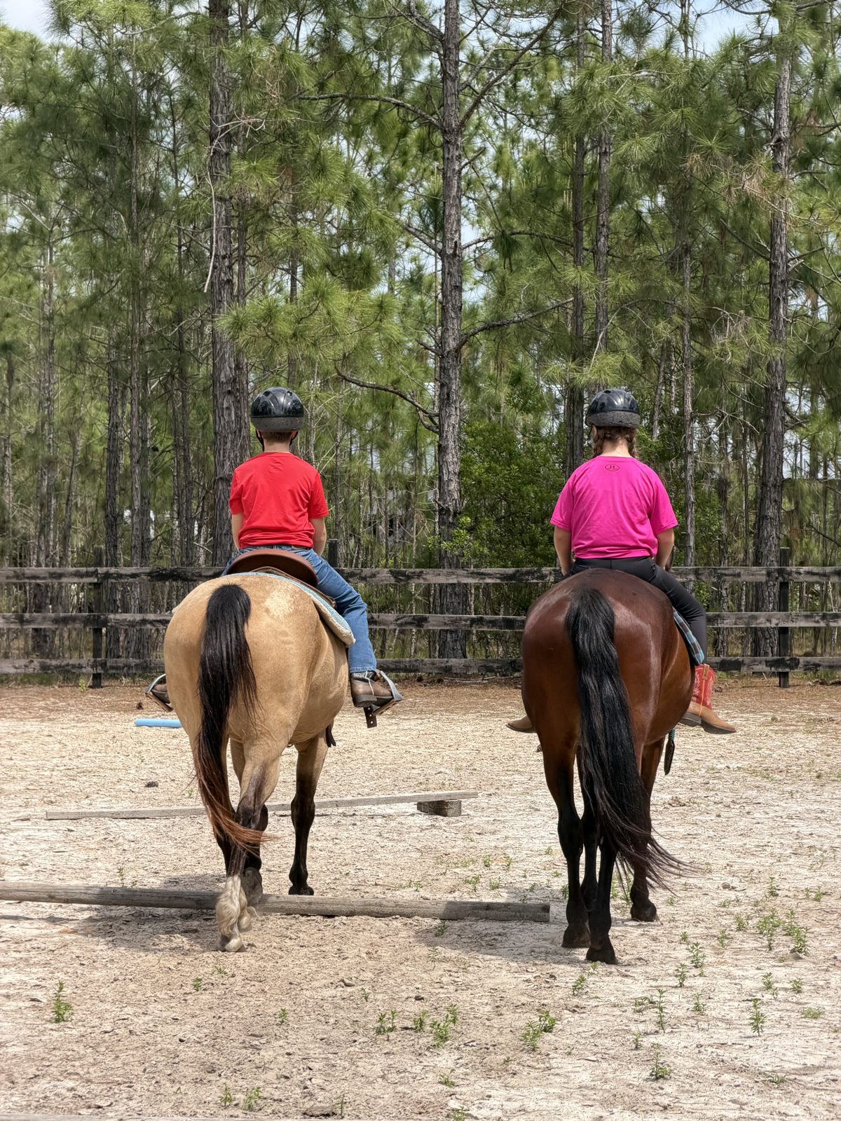 Two children ride side by side on a buckskin and a bay horse in the sandy arena at The Refuge
